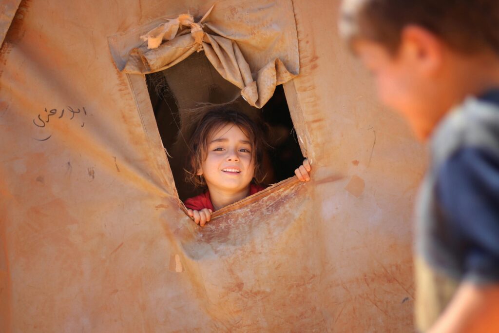 Positive ethnic girl looking out of window of tent located in refugee camp